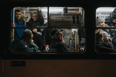 Inside a dimly lit public bus, a diverse group of passengers is visible through the window. People of various ages are engaged in different activities; some are seated, while others are standing. A man with a hat sits prominently in the center, appearing deep in thought, while nearby, passengers look outwards or converse. The atmosphere is crowded, indicating a busy commute.