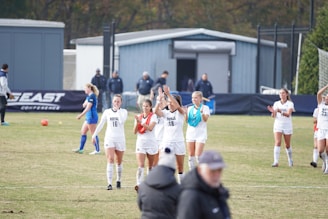A group of female soccer players wearing white uniforms are on a field, some clapping and others walking. In the background, players in blue uniforms, a sign with 'East Conference,' and several people are visible. The scene captures a moment of celebration or conclusion of a soccer match.