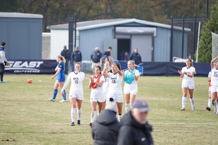 A group of female soccer players wearing white uniforms are on a field, some clapping and others walking. In the background, players in blue uniforms, a sign with 'East Conference,' and several people are visible. The scene captures a moment of celebration or conclusion of a soccer match.