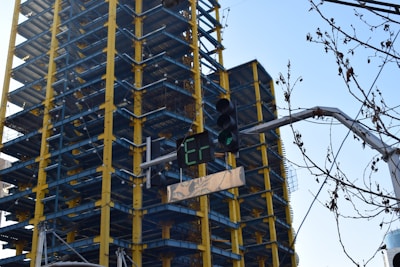 A multi-story building under construction with a visible steel framework painted in yellow and blue. In the foreground, a traffic light with green lights and a digital display is present. There are also some leafless tree branches in the scene.
