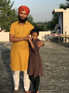 A man wearing an orange turban and yellow kurta stands on a rooftop with his arm around a young girl in a brown dress. They are smiling, and the background includes some greenery and a clear sky.