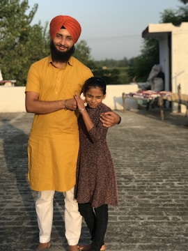 A man wearing an orange turban and yellow kurta stands on a rooftop with his arm around a young girl in a brown dress. They are smiling, and the background includes some greenery and a clear sky.
