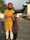A man wearing an orange turban and yellow kurta stands on a rooftop with his arm around a young girl in a brown dress. They are smiling, and the background includes some greenery and a clear sky.