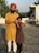 A man wearing an orange turban and yellow kurta stands on a rooftop with his arm around a young girl in a brown dress. They are smiling, and the background includes some greenery and a clear sky.