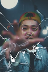 A person emerges from a blurry background, wearing a multicolored cap and denim jacket. The focus is on their face and hand, which is reaching out towards the viewer. The image appears fractured by cracks resembling broken glass, adding an abstract layer to the composition.
