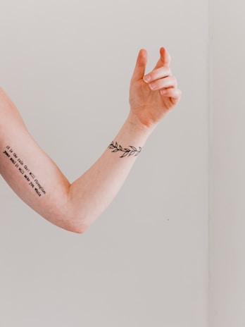 A forearm with a tattoo of a leaf design is raised against a plain background. The skin has a tattoo with text running along the inner forearm, adding an artistic and personal touch.