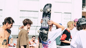 Several young people are gathered outdoors in a sunny environment. One person holds a skateboard above their head, wearing casual clothing. Others are shirtless or wearing relaxed attire, and there is a sense of social interaction and leisure.