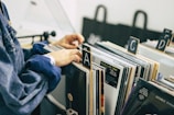 A person is sorting through a collection of vinyl records organized alphabetically in a store or library. The records are placed vertically in boxes, and the person's hand is flipping through them. The setting is calm, with muted colors, and the scene suggests a sense of nostalgia and appreciation for music.