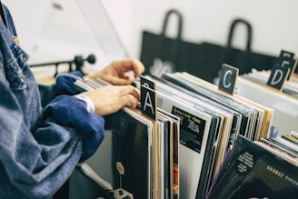 A person is sorting through a collection of vinyl records organized alphabetically in a store or library. The records are placed vertically in boxes, and the person's hand is flipping through them. The setting is calm, with muted colors, and the scene suggests a sense of nostalgia and appreciation for music.