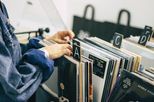 A person is sorting through a collection of vinyl records organized alphabetically in a store or library. The records are placed vertically in boxes, and the person's hand is flipping through them. The setting is calm, with muted colors, and the scene suggests a sense of nostalgia and appreciation for music.