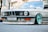A close-up view of the front end of a vintage BMW car, featuring a sleek design with prominent round headlights and a distinct front grille. The car sports custom turquoise rims, a lowered stance, and decals on the bumper. The surroundings suggest an urban setting with blurred background elements.