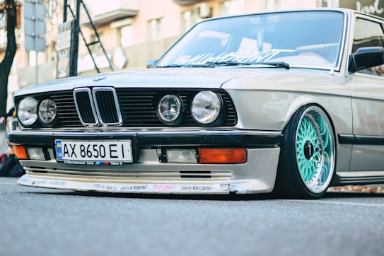 A close-up view of the front end of a vintage BMW car, featuring a sleek design with prominent round headlights and a distinct front grille. The car sports custom turquoise rims, a lowered stance, and decals on the bumper. The surroundings suggest an urban setting with blurred background elements.