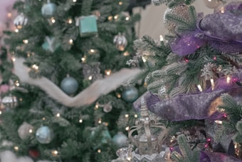 Two decorated Christmas trees with glowing lights and various ornaments including purple ribbons, blue and silver baubles, and sparkling silver decorations. The trees are lush with green pine branches and create a festive atmosphere.