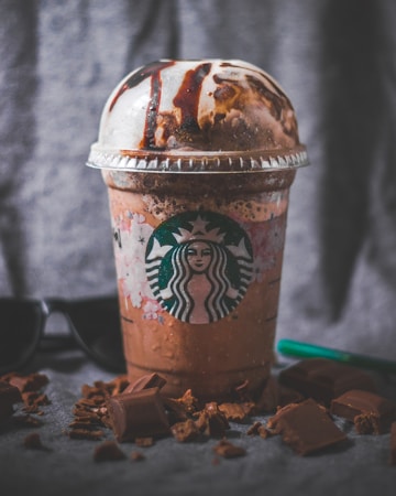 A creamy, chocolatey Starbucks frappuccino with chocolate drizzles on top is placed on a gray surface. Surrounding the cup are chunks of chocolate, creating a decadent and indulgent scene. A pair of dark sunglasses and a green straw rest nearby.
