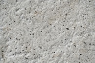 A close-up view of a textured concrete surface with small, irregular dark spots and a rough, uneven finish.