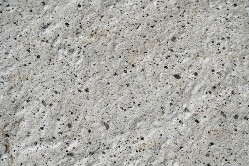 A close-up view of a textured concrete surface with small, irregular dark spots and a rough, uneven finish.