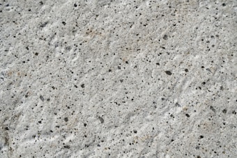 A close-up view of a textured concrete surface with small, irregular dark spots and a rough, uneven finish.