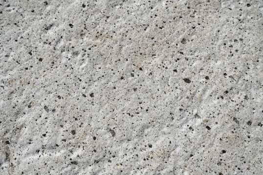 A close-up view of a textured concrete surface with small, irregular dark spots and a rough, uneven finish.