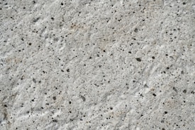 A close-up view of a textured concrete surface with small, irregular dark spots and a rough, uneven finish.