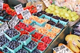 A colorful display of fresh berries and fruits is arranged in small blue cartons. The assortment includes blueberries, strawberries, blackberries, and raspberries, all labeled with price tags. Behind the berries, a selection of green and yellow fruits like lemons and limes is visible.