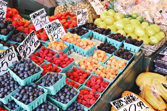 A colorful display of fresh berries and fruits is arranged in small blue cartons. The assortment includes blueberries, strawberries, blackberries, and raspberries, all labeled with price tags. Behind the berries, a selection of green and yellow fruits like lemons and limes is visible.