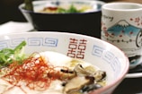 A close-up view of a bowl of ramen, featuring sliced mushrooms, green onions, a soft-boiled egg, and thin red chili strands. The bowl has blue geometric patterns and red characters on the side. In the background, a decorative cup with illustrations of Mount Fuji and cherry blossoms is partially visible.