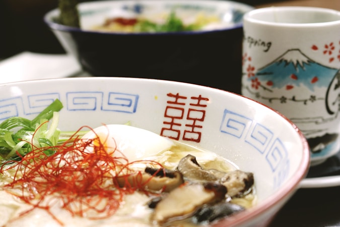 A close-up view of a bowl of ramen, featuring sliced mushrooms, green onions, a soft-boiled egg, and thin red chili strands. The bowl has blue geometric patterns and red characters on the side. In the background, a decorative cup with illustrations of Mount Fuji and cherry blossoms is partially visible.