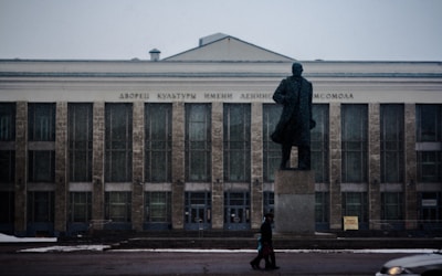 A large, imposing building with multiple windows and a sign in Cyrillic text above the entrance. In front of the building stands a statue of a man on a pedestal, silhouetted against the structure. Snow is lightly falling, and a person walks past the monument carrying a bag. A white car is partially visible on the right side of the image.