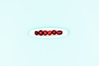 Seven red cranberries are neatly arranged in a small, elongated white dish set against a light blue background. The minimalistic composition emphasizes the vibrant color of the cranberries in contrast to the soft pastel backdrop.