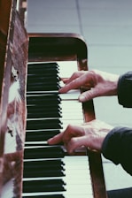 Hands playing a piano, focusing on the keys and fingers in action. The wood of the piano and the contrast between the black and white keys create a classic, artistic atmosphere.