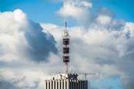 A telecommunications tower is mounted on top of a high-rise building. The sky is filled with large, fluffy clouds, giving a dynamic and textured backdrop.