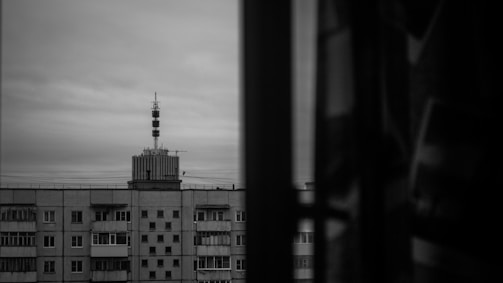 A grayscale image depicting a cityscape. The main focus is a mid-rise apartment building with a rooftop tower featuring multiple antennas. The building has numerous windows and balconies, and the sky is overcast. The right side of the image shows a dark, blurred vertical element, possibly a window frame or part of an interior.