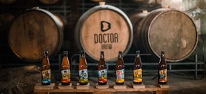 Several beer bottles with colorful labels are displayed in front of large wooden barrels in a dimly lit brewery setting.