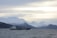 A cargo ship sails on a cold, choppy sea with snow-capped mountains in the background under a cloudy sky.