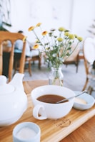A cozy café scene with a wooden table featuring a cup of tea, a white teapot, and small bowls containing sugar and a spoon. A vase of yellow flowers adds a touch of color, and blurred chairs and tables are visible in the background.
