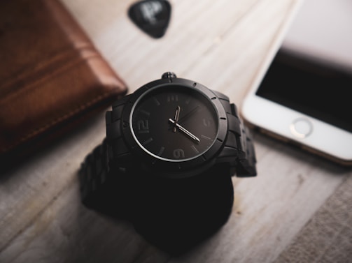 A sleek watch resting on a wooden table beside a folded pant and leather shoes.