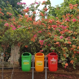 Three colorful waste bins are placed in front of a vibrant garden filled with pink and orange flowering shrubs. The bins have different labels indicating their use for different waste types. The background is lush with greenery and bright flowers, creating a lively contrast.