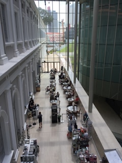 The image captures a modern indoor space with a high ceiling and large glass windows, allowing natural light to illuminate the area. People are seated at round tables, possibly in a cafe or common area, engaging in conversation. The interior design features sleek lines and a minimalistic style, with a blend of white and glass elements. A bookshelf or display stand containing various items is visible along one side.