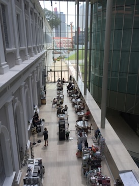 The image captures a modern indoor space with a high ceiling and large glass windows, allowing natural light to illuminate the area. People are seated at round tables, possibly in a cafe or common area, engaging in conversation. The interior design features sleek lines and a minimalistic style, with a blend of white and glass elements. A bookshelf or display stand containing various items is visible along one side.