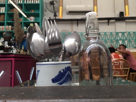 A dining table is set with a small ceramic cup holding silverware including spoons and forks. Next to it is a clear glass bottle with a swing top cap. In the background, there is a teal-colored shelving unit filled with various items and a couple of diners sitting at a table with rattan chairs. The decor features modern and eclectic elements.