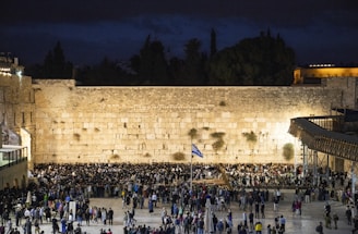 A large gathering of people stand in front of an ancient limestone wall, illuminated at night. The scene is lively with a mix of individuals, some in traditional attire. A flag is visible in the foreground, and the background is a mix of dark sky and structures.