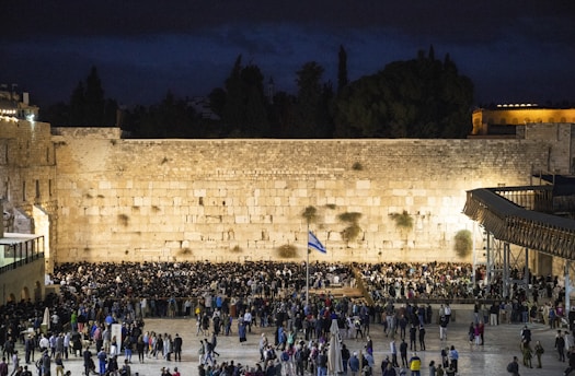 A large gathering of people stand in front of an ancient limestone wall, illuminated at night. The scene is lively with a mix of individuals, some in traditional attire. A flag is visible in the foreground, and the background is a mix of dark sky and structures.