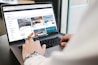 A person is using a laptop to browse an online marketplace featuring various digital products or themes. The screen displays a selection of software or website themes with descriptions and prices. The person seems to be in a well-lit room, sitting at a table.