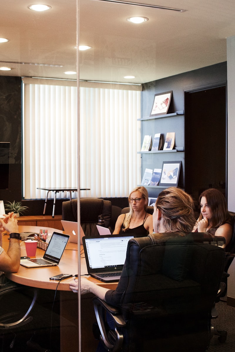 Image is a boardroom with 3 women sitting around a table in a meeting