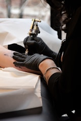 A tattoo artist is in the process of creating a tattoo on a person's arm using a tattoo machine. The artist is wearing black gloves and has a bracelet on their wrist. The setting appears to be a tattoo studio with disposable materials placed under the arm.