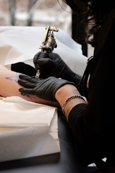 A tattoo artist is in the process of creating a tattoo on a person's arm using a tattoo machine. The artist is wearing black gloves and has a bracelet on their wrist. The setting appears to be a tattoo studio with disposable materials placed under the arm.