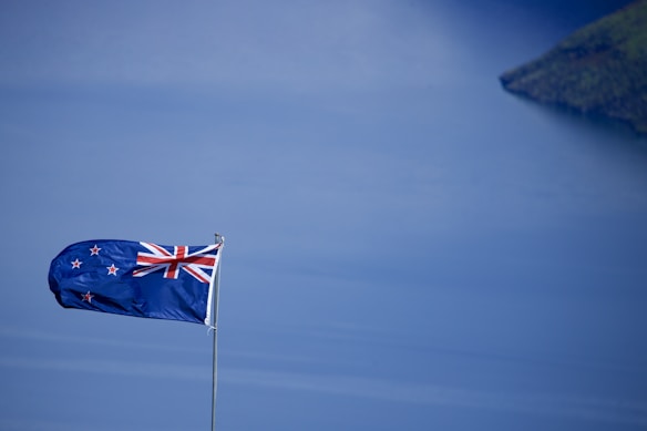 A New Zealand flag waves prominently against a serene backdrop of a vast, blue body of water. The sky above mirrors the water&rsquo;s tranquil shades, creating a harmonious scene. A distant piece of land peeks from the top right corner, adding depth to the image.