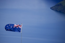 A New Zealand flag waves prominently against a serene backdrop of a vast, blue body of water. The sky above mirrors the water&rsquo;s tranquil shades, creating a harmonious scene. A distant piece of land peeks from the top right corner, adding depth to the image.