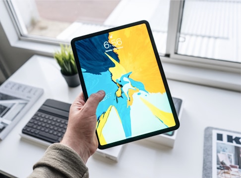 A hand holding a tablet with a vibrant abstract artwork displayed on the screen. The background shows a white desk with a keyboard, a small potted plant, and some magazines. Natural light is coming in from the window, creating a bright and modern atmosphere.