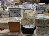 Various types of cheese are displayed on a counter in a market or store. Each cheese has a label with its name, origin, and price. The cheeses vary in color and texture, and the signs indicate different types of milk sources and flavor profiles.